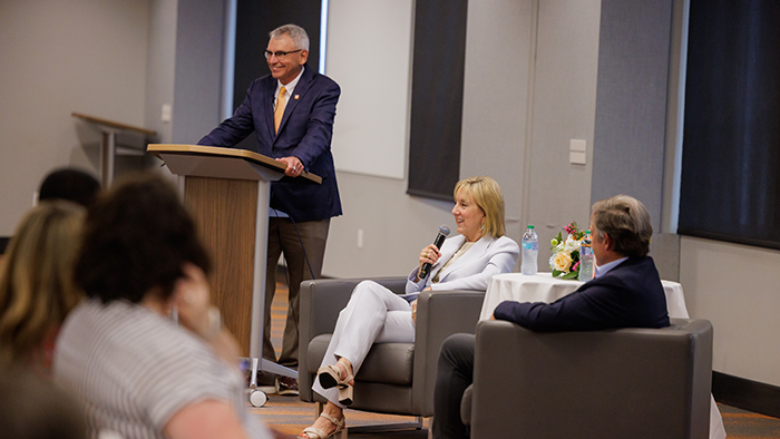 A man and woman in suits sit while talking to a room of people, with another man moderating from behind a podium.