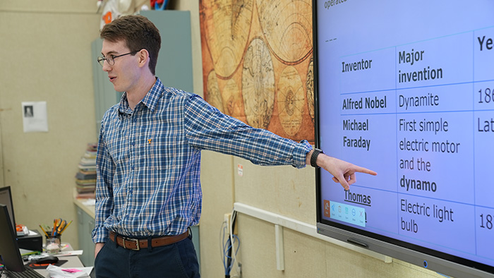 A man with glasses, wearing a plaid shirt, points to a presentation screen with teaching a class.