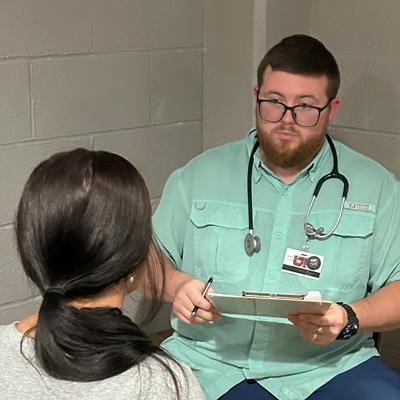 A man with glasses, wearing a green fishing shirt and stethoscope, talks with a patient.