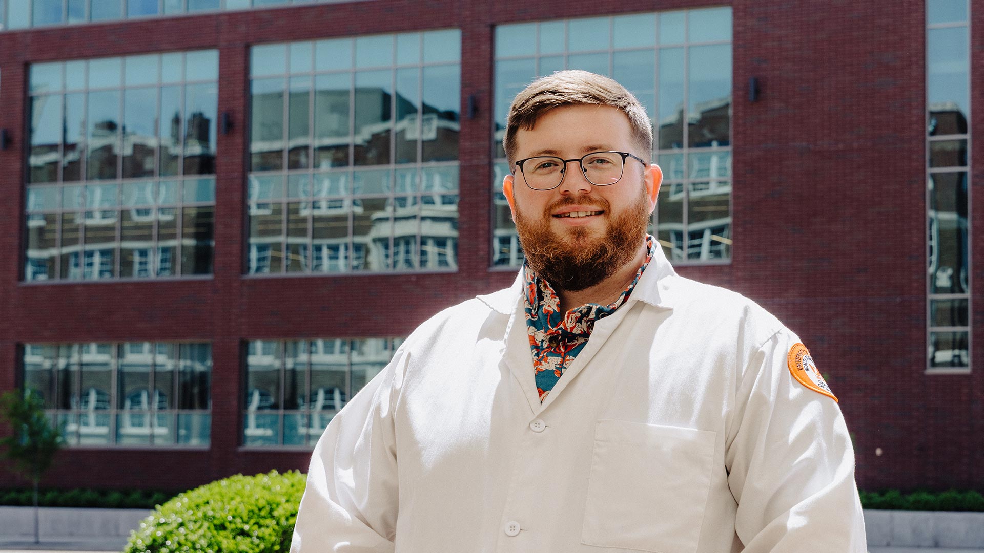 A bearded man with glasses, wearing a white medical coat, standing outside in front of a building.