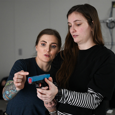 A woman instructs a female student on the operation of a simulator hand gun.