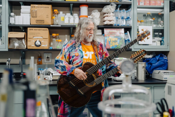 An older man with long hair and beard, wearing a tie-dye lab coat, plays a double-neck bass guitar while in his laboratory.