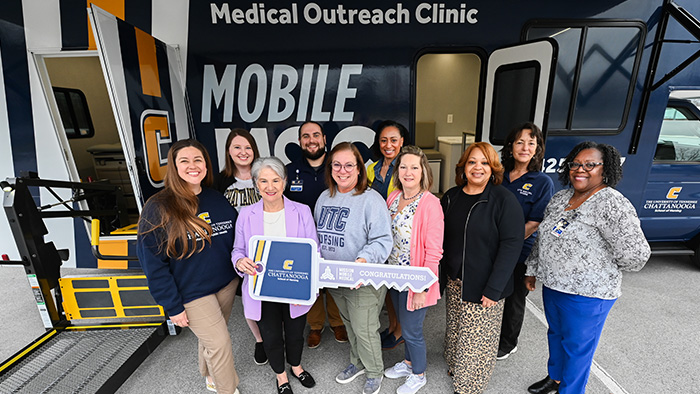 A group of people holding a ceremonial key, while standing in front of a blue, white and gold mobile health clinic.