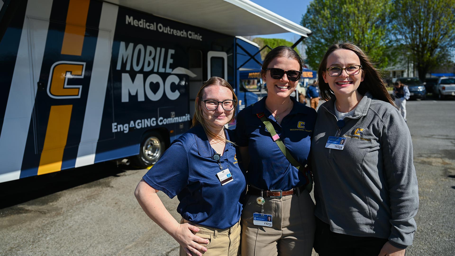 Three young women standing together for a photo in front of a blue, white and gold mobile health clinic.