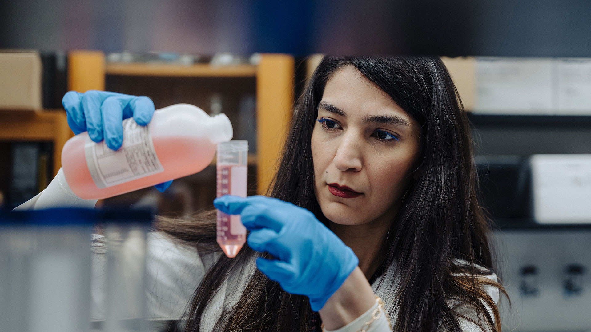 A female scientist carefully pours a pink liquid into a clear container with measurement markings.