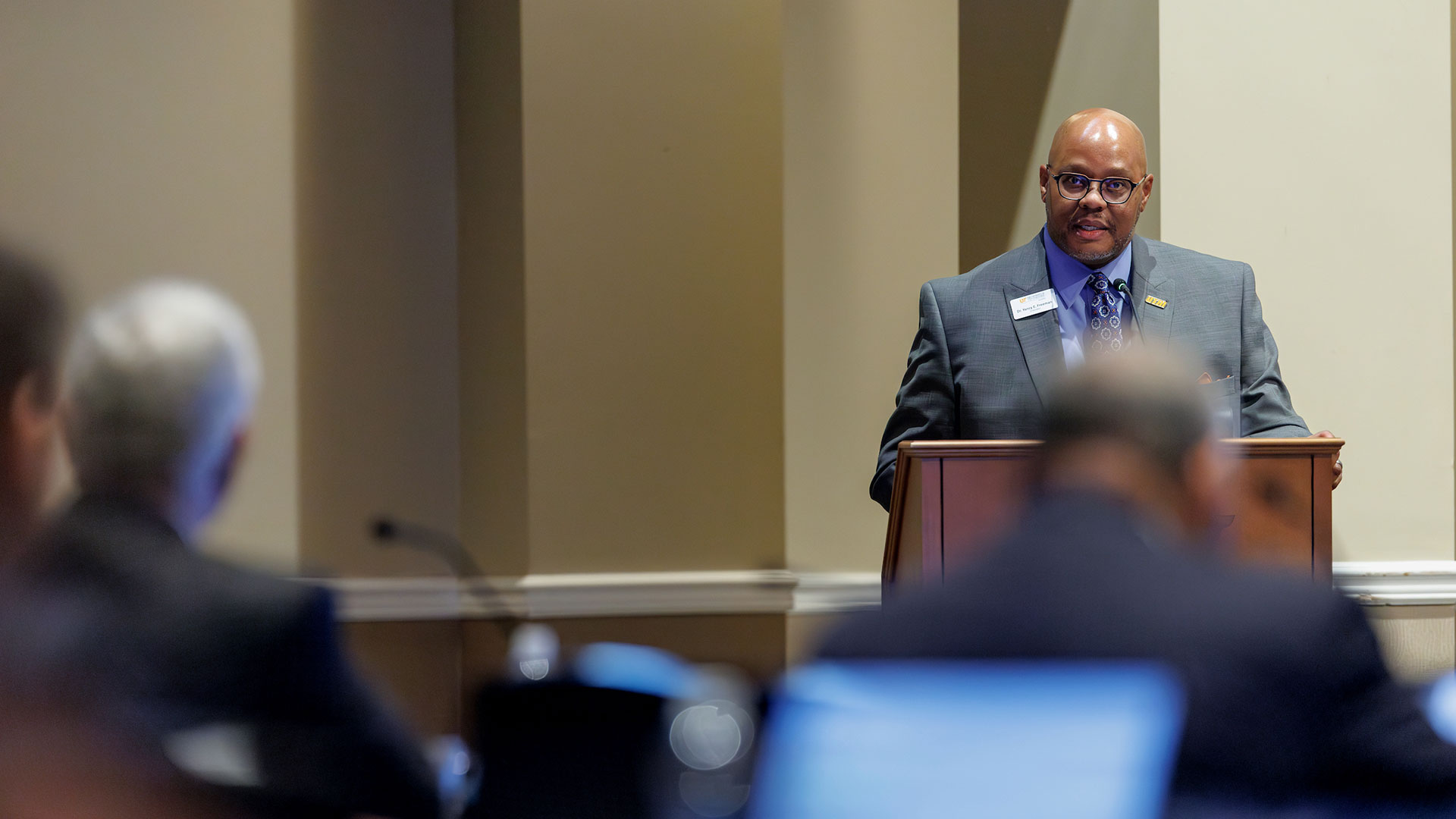 A black male wearing a blue and gray suit speaks to room from behind a podium.