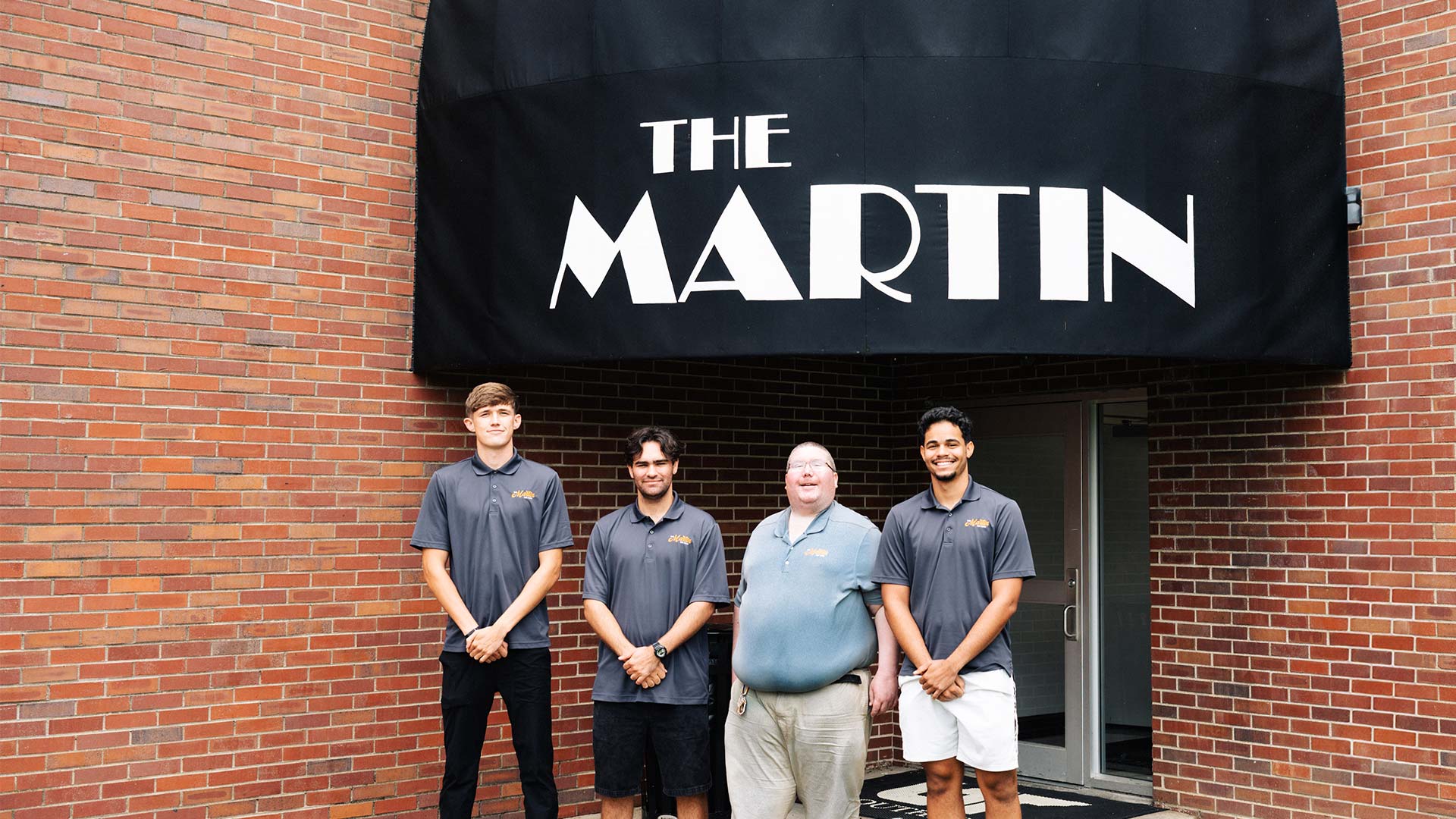 Four young men wearing gray polo shirts standing in front of a red brick building, with a black entrance covering that says "The Martin."