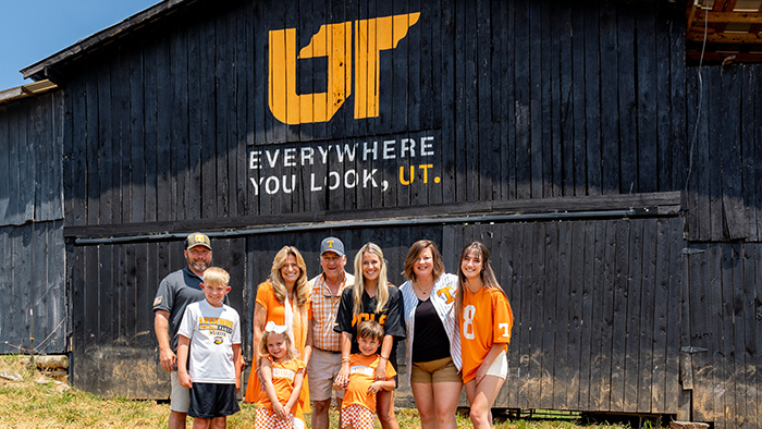 A family dressed in Tennessee orange and white stand in front of a old, black barn with the 'Everywhere You Look, UT' logo painted on it.