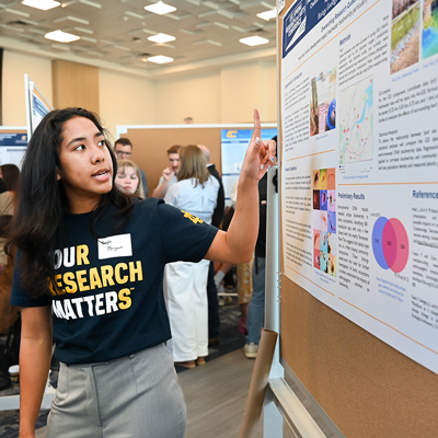 A female student points at a board while presenting a research poster.