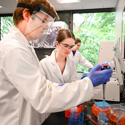 Three students wearing lab coats and safety equipment working in a college laboratory.