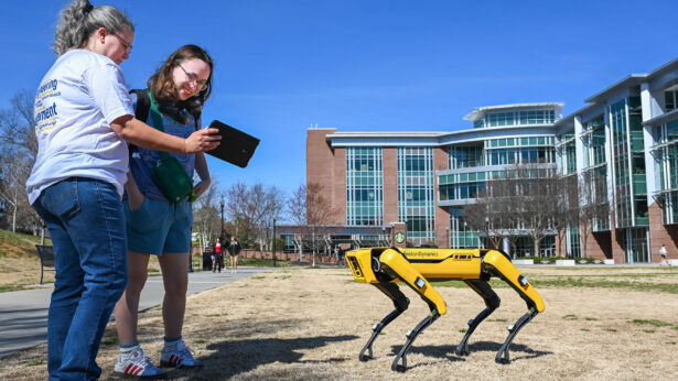 A student and professor examine data gathered from a robotic dog, outside on a grassy field.