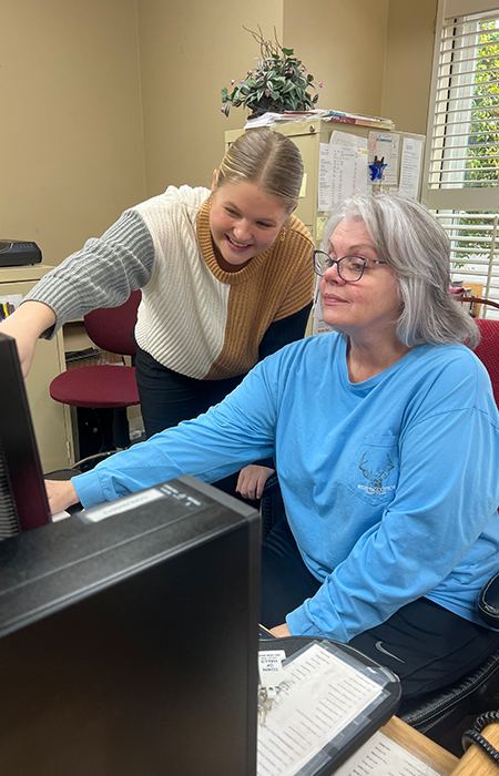 A young blonde woman pointing at a computer screen while an older woman sits and operates the computer.