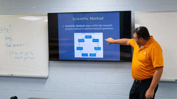 A man in an orange polo shirt points at a presentation screen during a class.