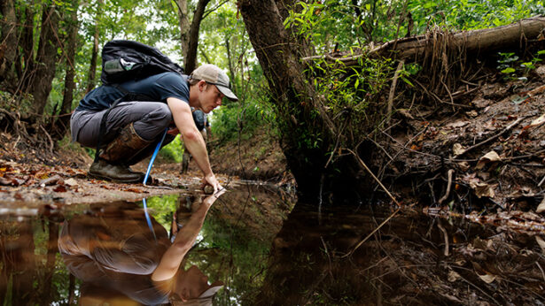 A young man in outdoor gear crouches down in a forest, examining a stream for wildlife.