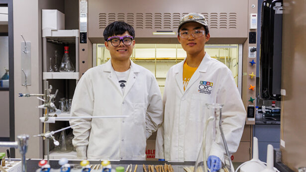 Two asian students wearing lab coats pose for a photo while in their lab.