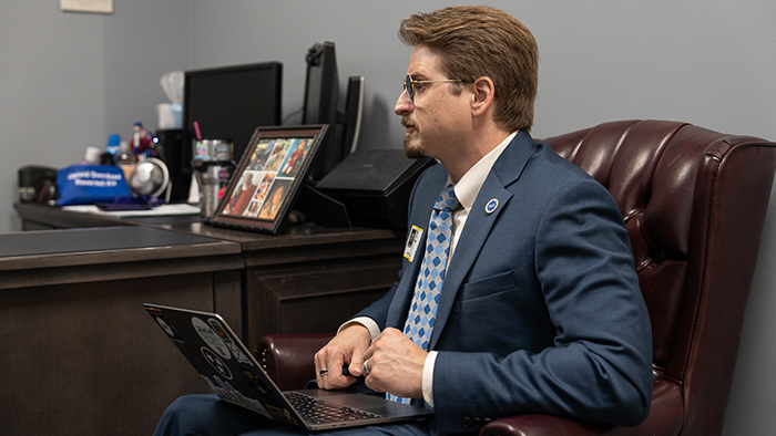 A man in business suit, sits in a office while working with a computer in his lap.
