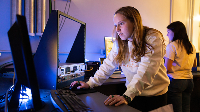 A female student leans over a desk while operating a computer and examining the display.