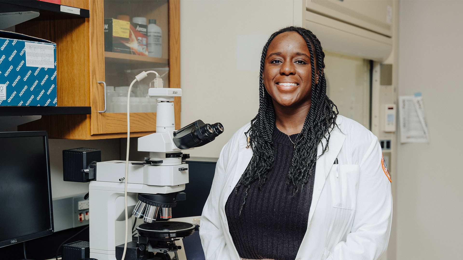 A black woman wearing a white lab coat while standing next to a microscope.