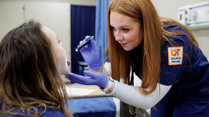 A redheaded student in nursing scrubs uses a penlight to examine a patient's mouth.