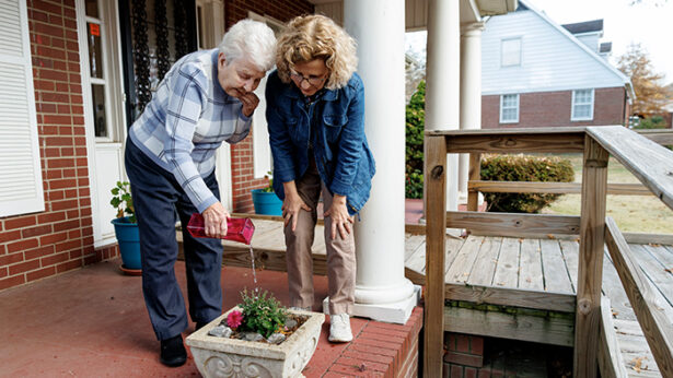 Two women watering a plant on an outside brick porch.