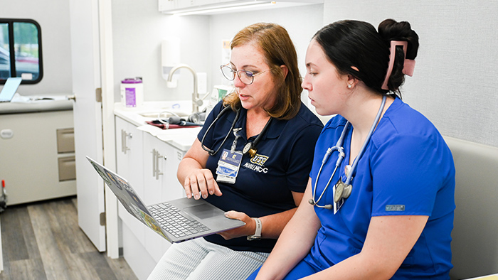 Two women sitting in white health clinic room look at a laptop.