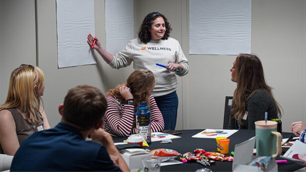 A woman speaks to a group while pointing to a white board.