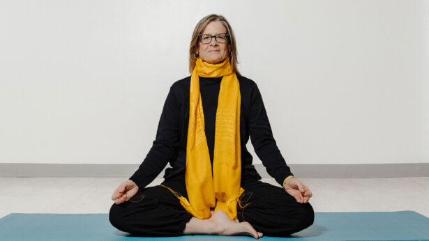 A woman in black outfit and yellow scarf, sitting in a cross-legged yoga pose.