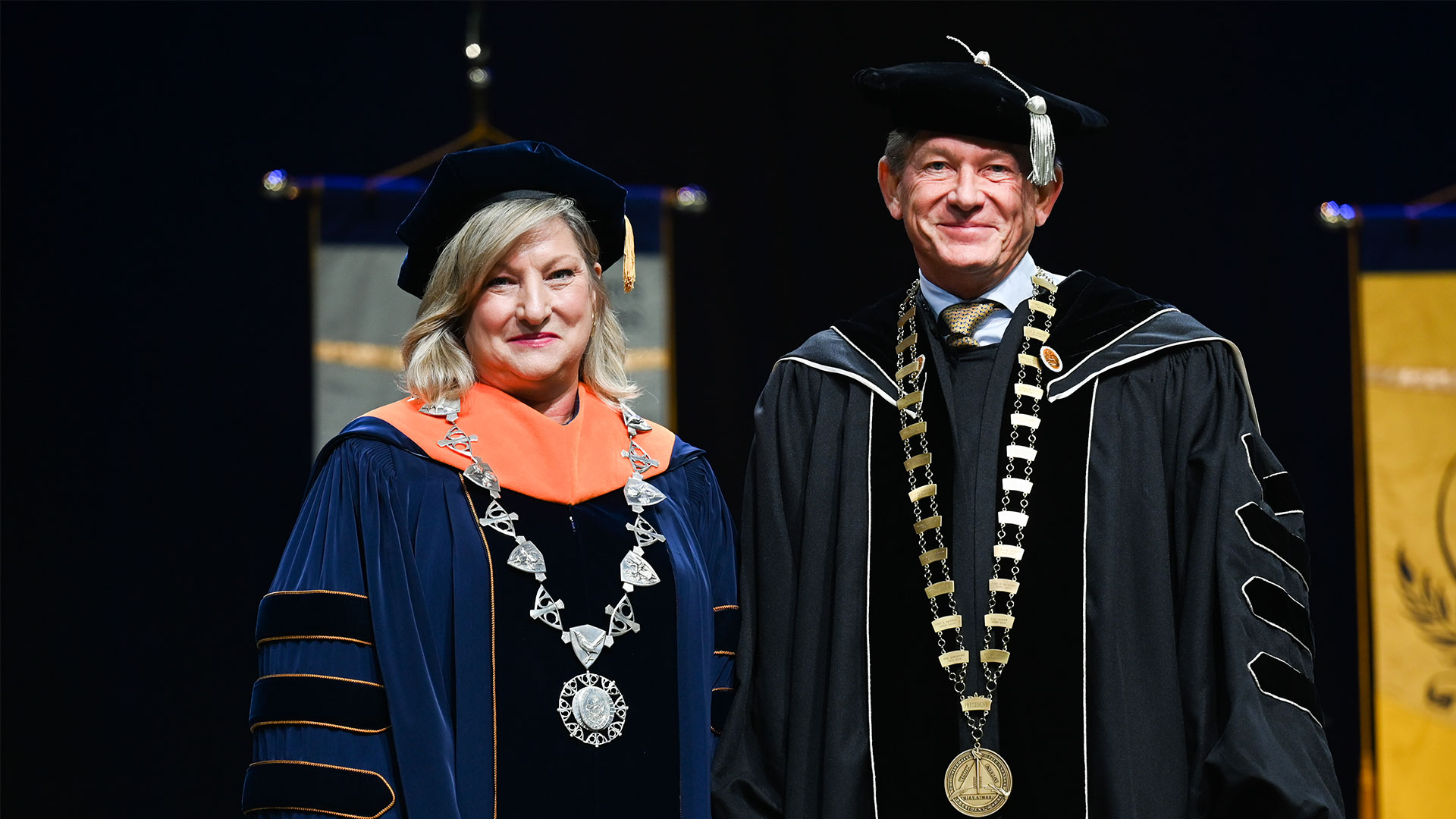 A man and woman standing side-by-side, both wearing graduation regalia.