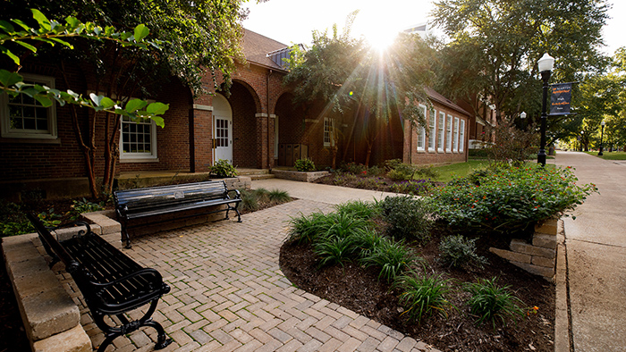 Exterior brick walkway of a red brick building, surrounded by plants and shrubbery.