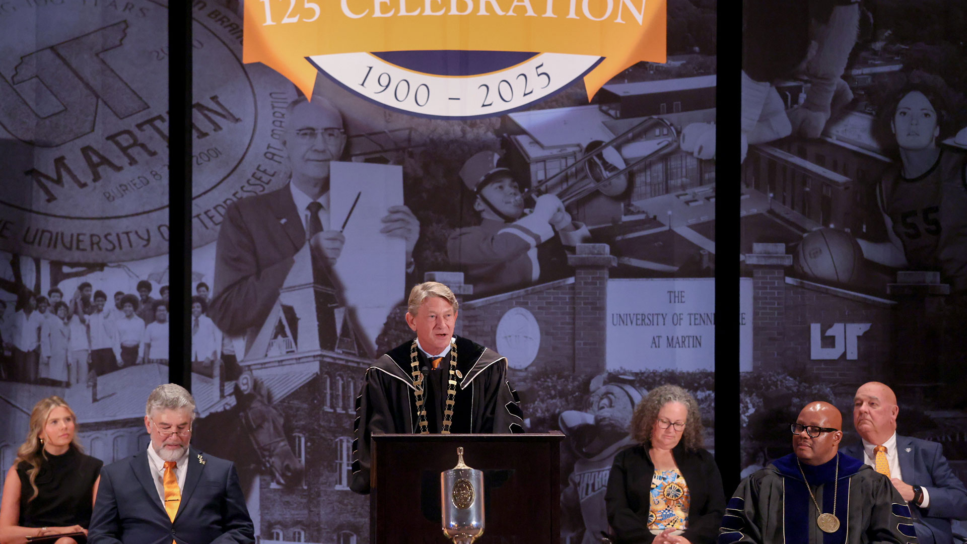 A man in graduation regalia speaks from behind a podium, with a historical themed digital display behind him.
