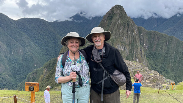 A man and woman posing for a photo mid-hike, with mountains and clouds in the background.