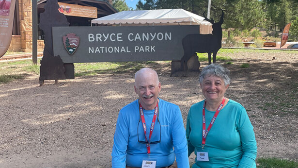A man and woman sitting in front of a wooden sign that says "Bryce Canyon National Park."