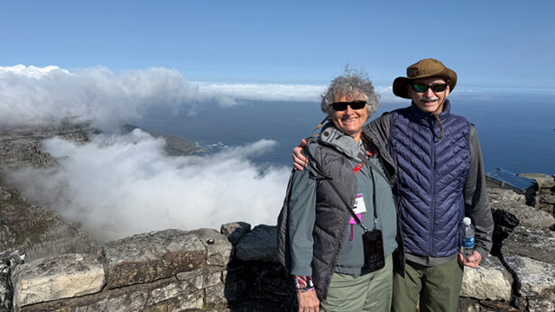 A man and woman posing for a photo, with the ocean and clouds in the background.