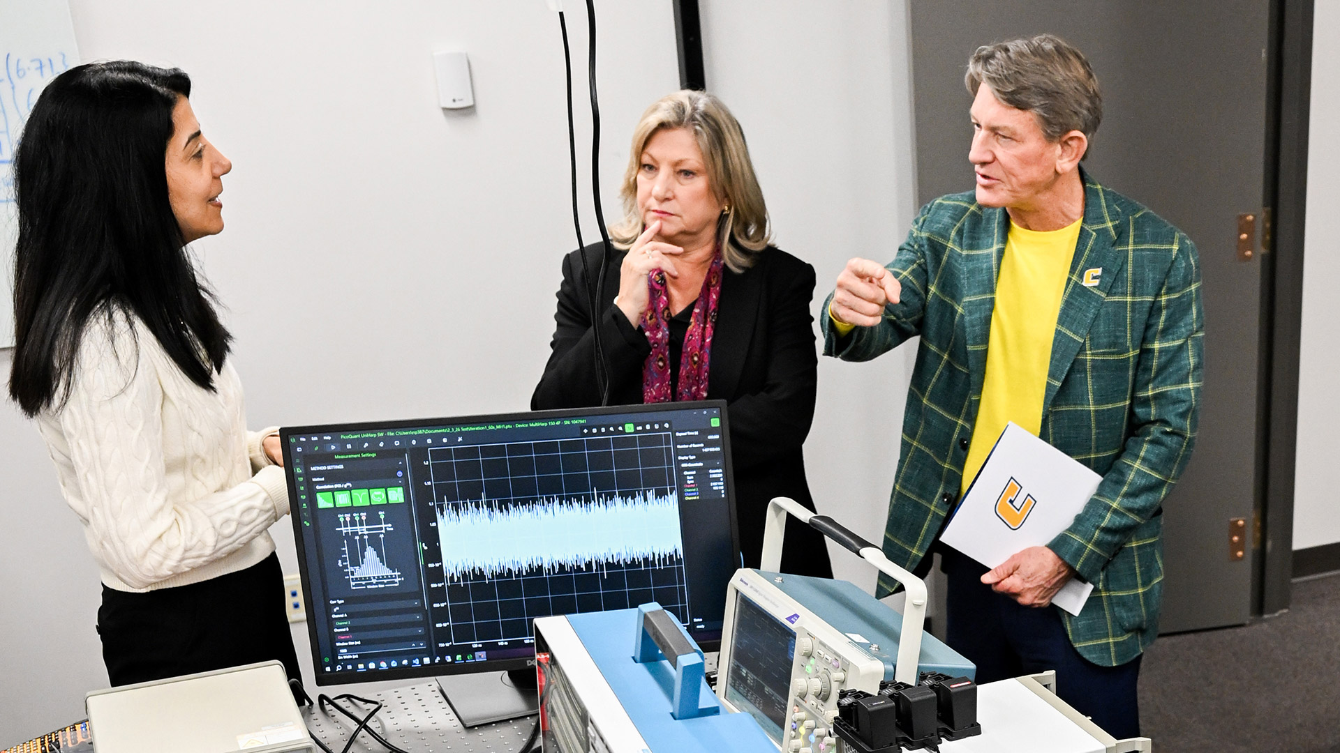 A man and two women talking next to a computer with research read-out displays.