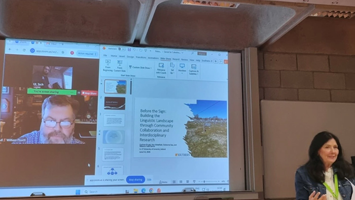 A woman delivering a presentation, with virtual participants on display in the background.