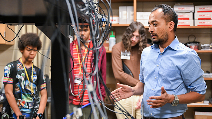 A group of people listen to a man talking, with a table of equipment and wires in front of them.