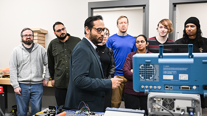 A group of people listen to a man talking, with a table of equipment in front of them.
