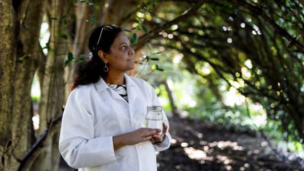 A woman in white lab coat, standing under trees while holding a jar filled with water.