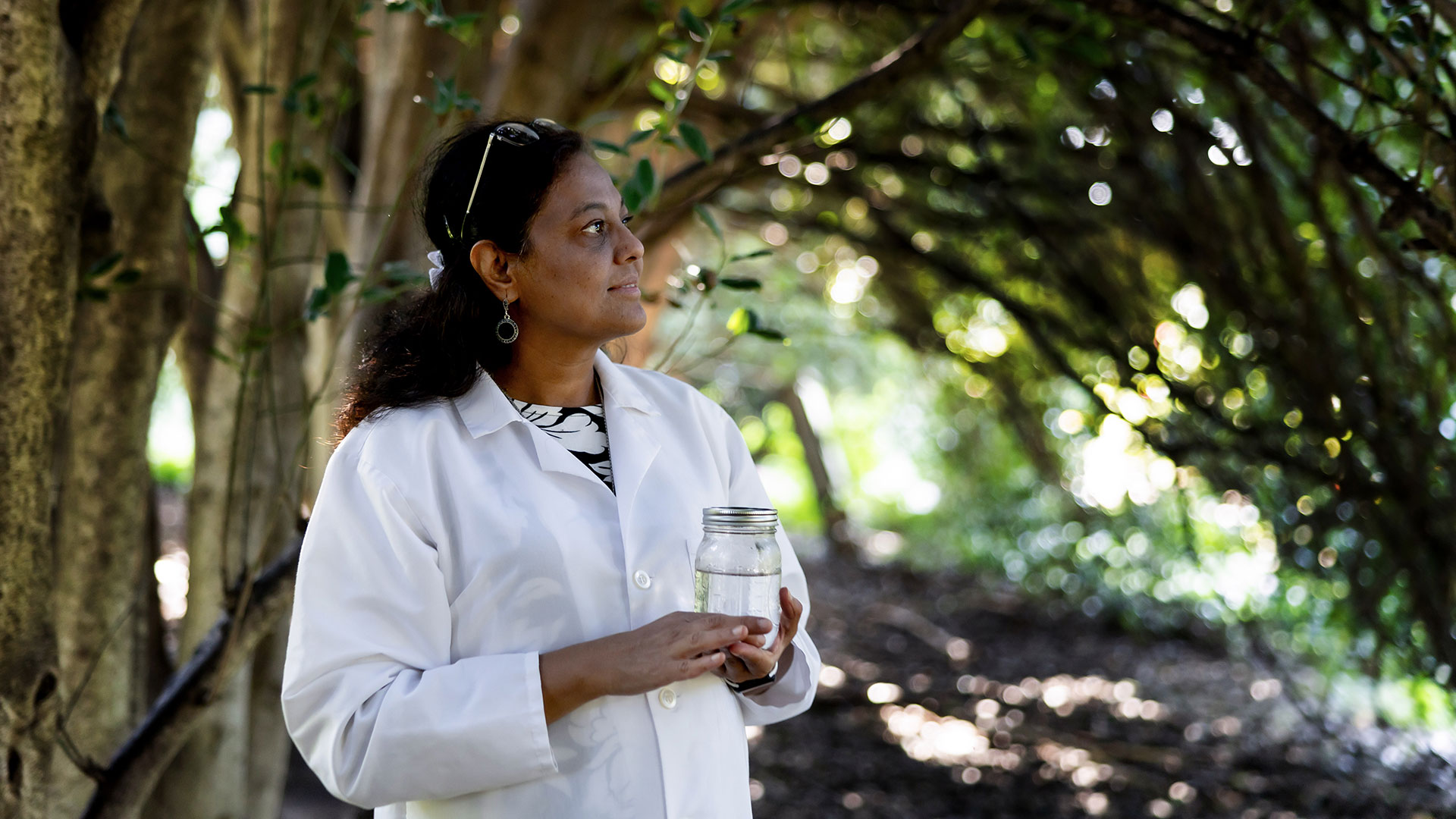 A woman in white lab coat, standing under trees while holding a jar filled with water.