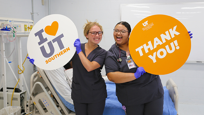 Two female nurses in gray scrubs smile while holding up "thank you" signs.